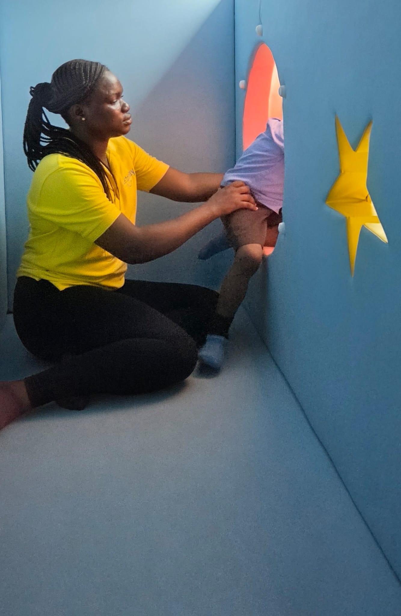 A Letzplay staff member in yellow gently guiding a toddler through a porthole in the soft play wall.